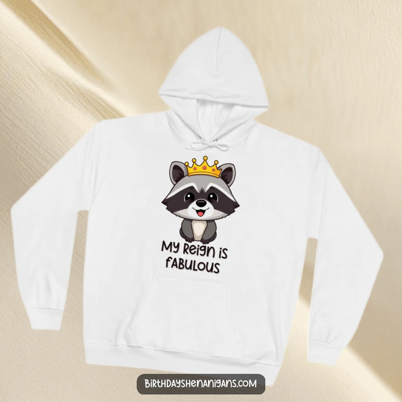 Funny hoodie depicting a happy raccoon character wearing a glittering, festive crown, embodying playful royalty and cozy vibes.